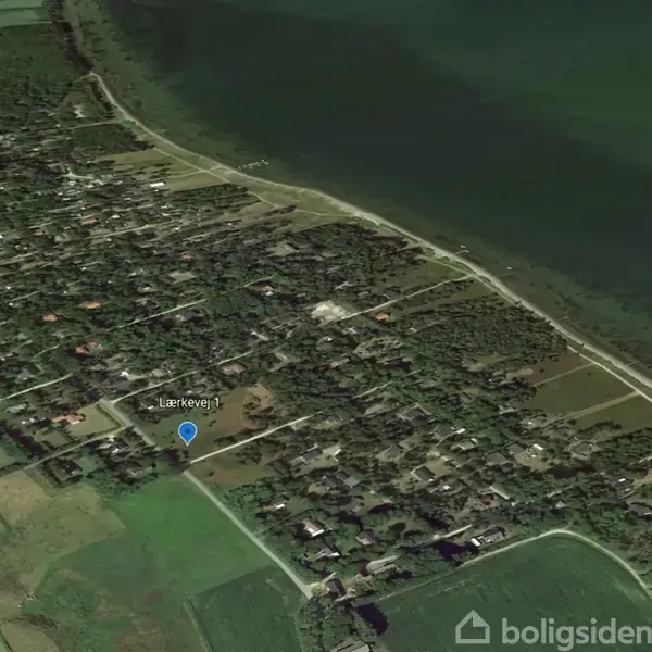 Satellitbillede viser et område ved kystlinjen med mange huse og veje omgivet af grønne marker og skov. En blå markør indikerer "Lærkevej" nær et beboelsesområde.