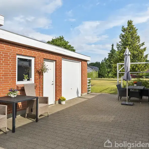 Rød murstensbygning med udendørs terrasse inkluderer bord og stol med plante. I baggrunden ses grøn have og træer med spisebord og parasol. Solrigt med blå himmel.