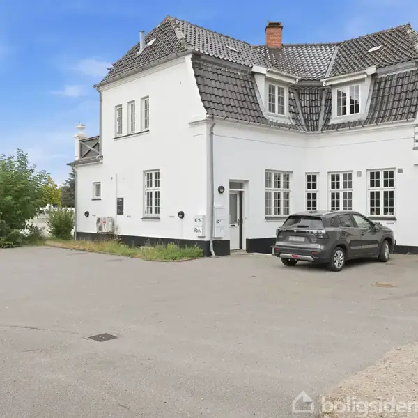 Hvid to-etagers bygning med skråt tag og kviste, parkeret bil foran. Omkringliggende asfaltområde med græsklædte kanter, blå himmel i baggrunden.