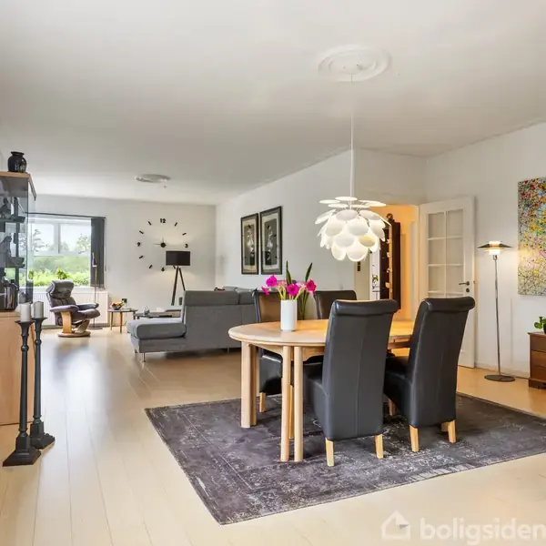 Rundt spisebord med fire sorte stole står på et tæppe i en stue. Baggrunden har en sofa, farverige malerier, en stor vindue og en reol. Stor loftslampe hænger over bordet.