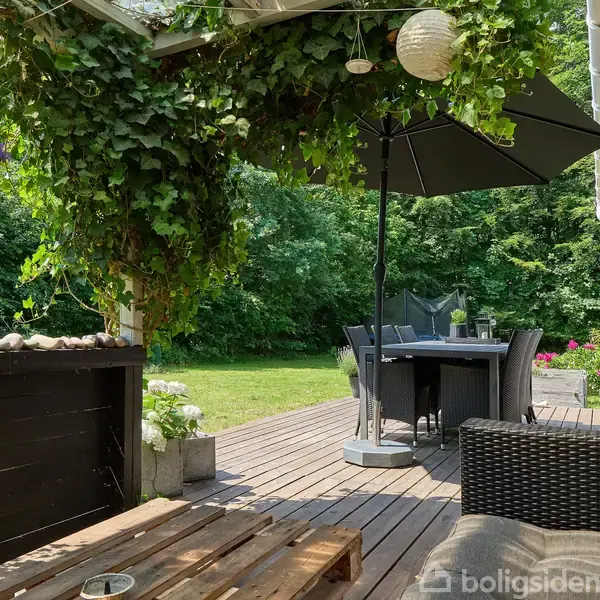Terrasse med træbord og -stole, overdækket af grønne planter, under åben parasol. I baggrunden en græsplæne og træer. Atmosfære af sommerhusidyl og afslapning i naturen.