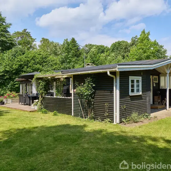 Sort træhus står stille omgivet af grønt græs og frodige træer. Sollys skinner på huset, der har et fladt tag og små vinduer. Atmosfæren er fredelig og sommerlig.