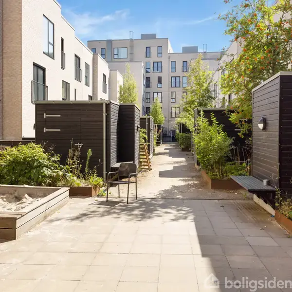 Sandkasse står stille i et gårdmiljø omgivet af murstensbygninger og grønne planter. Sollys belyser fliseområdet, hvor små skure og bænke ses.
