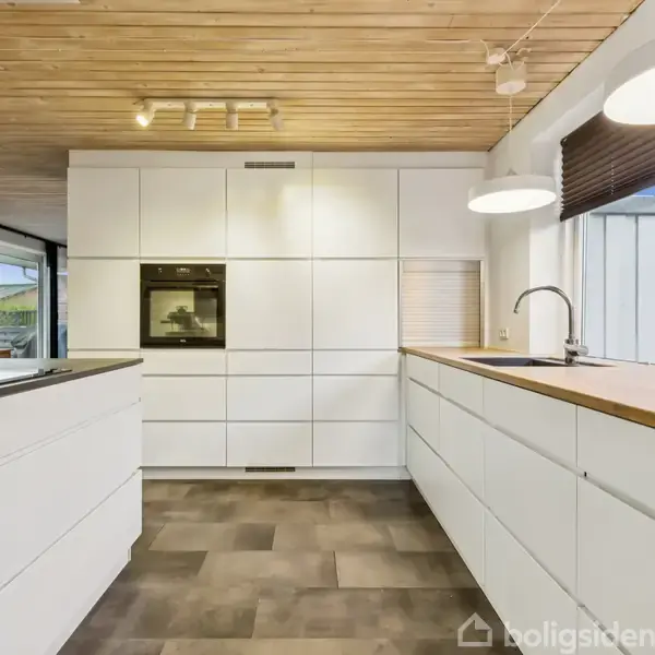 Et moderne køkken med hvide skabe og indbygget ovn. En køkkenø med sort bordplade står centralt. Træloft og lyse lamper skaber en varm atmosfære.