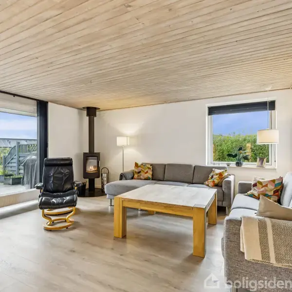 Stue indeholder en grå hjørnesofa med farverige puder, et sort læderarmatur, og et træbord på lys trægulv. Et stort vindue og en dør til venstre leder ud til en terrasse.