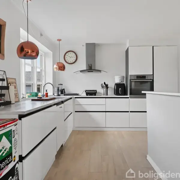 Et moderne køkken med hvide skabe og en sort bordplade; på væggen hænger en tavle med teksten "HYGGE i KØKKENET". I baggrunden ses kobberlamper og en vægur.