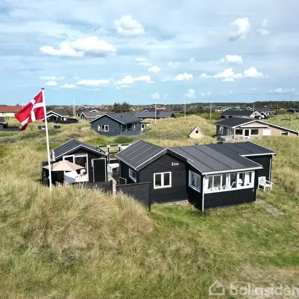 Sort sommerhus står stille omgivet af græsbevoksede klitter og andre huse; et dansk flag vejrer i vinden. Teksten "E209" ses på huset.