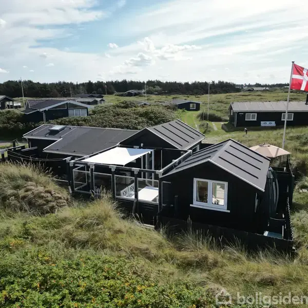 Sort hus med flere bygningsafsnit står stille på en sandet og græsbevokset grund, omgivet af andre lignende huse. Dannebrog vajer på en stang i baggrunden under en blå himmel.
