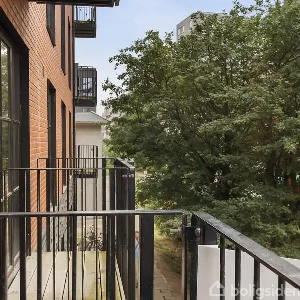 En balkon med sort metalgelænder er forbundet til en murstensbygning. Den vender ud mod et grønt træ, der står i nærheden, med flere bygninger i baggrunden.