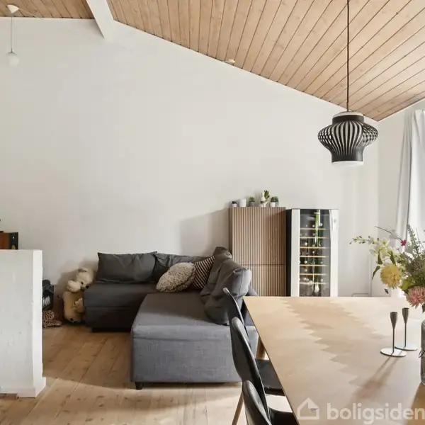 Gråt sofaarrangement står i en lys stue med skråt træloft. Ved siden af er et spisebord med blomster. Store vinduer lader lys ind, og to sorte lamper hænger ned.