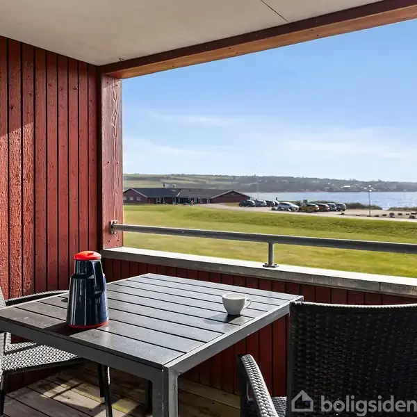 En terrasse med et bord og to stole, hvor en termokande og kop står. Udsigt over en græsplæne og vand i baggrunden under en blå himmel.