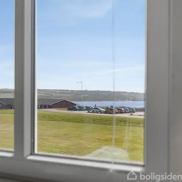 Udsigt gennem et vindue viser parkerede biler nær en sø med græsklædt område i forgrunden og et bakkeområde i baggrunden under en klar blå himmel.