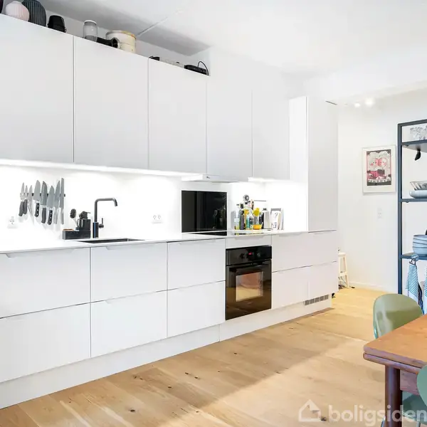 Et moderne køkken med hvide skabe, en sort ovn og indbygget kogeplade. En række knive hænger på væggen over vasken. Trægulv og spisebord til højre i billedet.