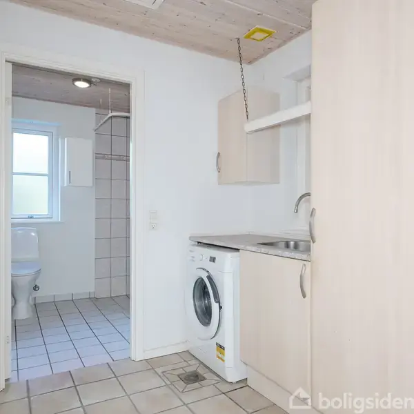 Vaskemaskine står under en vask i et lyst bryggers med fliser og træloft; dør leder til et flisebelagt badeværelse med toilet og vindue.