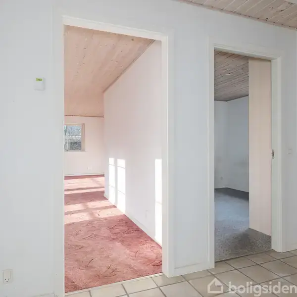 Et indendørs rum med to døre, der fører til separate værelser. Et rum har et rødt gulvtæppe og sollys fra et vindue. Væggene er hvide, gulvet flisebelagt.