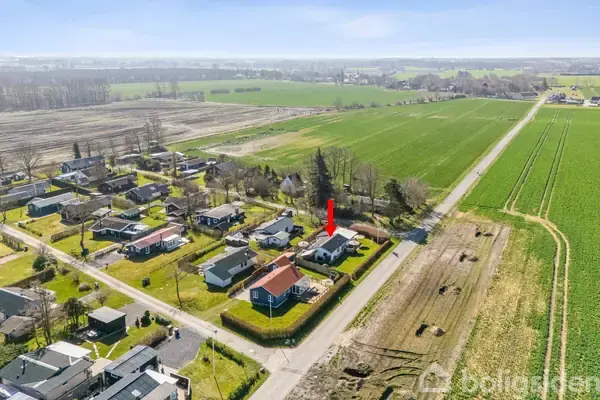 Huse ligger spredt i et landligt område. En rød pil peger på et specifikt hus blandt de grønne marker og andre hjem. Det er solrigt, og landskabet er fladt.