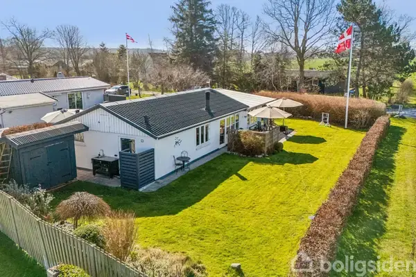 Et hvidt hus med mørkt tag står i en velplejet have med grøn græsplæne. To flagstænger med danske flag er placeret omkring huset, omgivet af buske og træer.