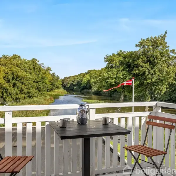To træstole og et bord står på en terrasse med hvidt rækværk. Kaffe i en stempelkande står på bordet. Baggrunden viser en flod og skovklædte omgivelser med en rød flag.
