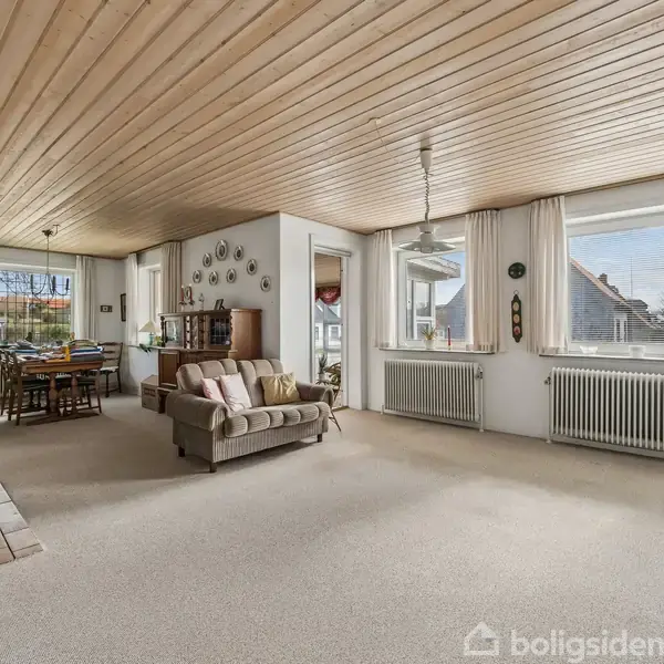 Stue med beige sofa, sofabord og spisebord i baggrunden. Træloft og lyse vægge, store vinduer med dagslys. Radiatorer under vinduerne, spredte dekorationer på væggene.