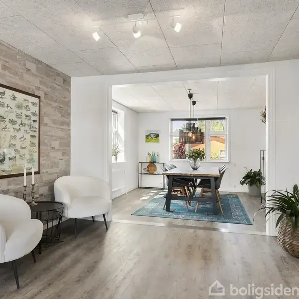 To lænestole står ved siden af et lille bord i en opholdsstue. På væggen er en plakat, og gennem en åbning ses et spisebord med stole. Planter pynter rummet.