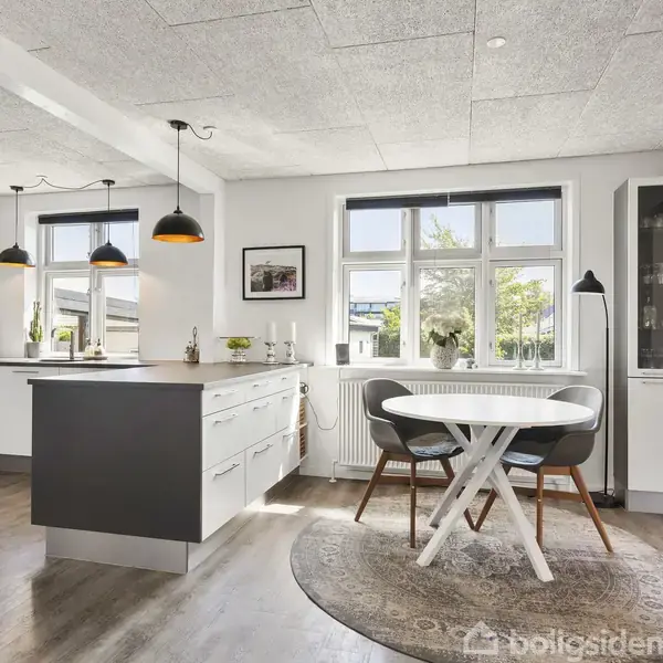 Køkken og spiseområde med mørkke køkkenskabe og hvide skuffer, en rund spisebord med stole på gulvtæppe, store vinduer med udsigt og moderne lamper i et lyst rum.