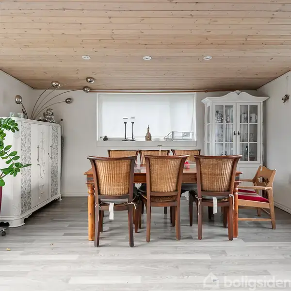 Spisebord med seks stole står midt i et rum med lyst trægulv og træloft. Til venstre en stor plante, til højre en glasskab. Sollys strømmer ind fra store vinduer.