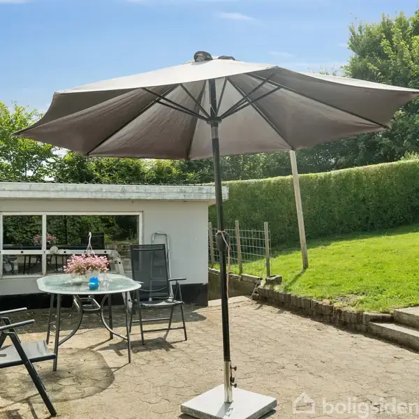 En stor parasol står på en terrasse med et havebord og to stole. Omkring er der grønne buske og et græsareal, mens en lav bygning står i baggrunden.