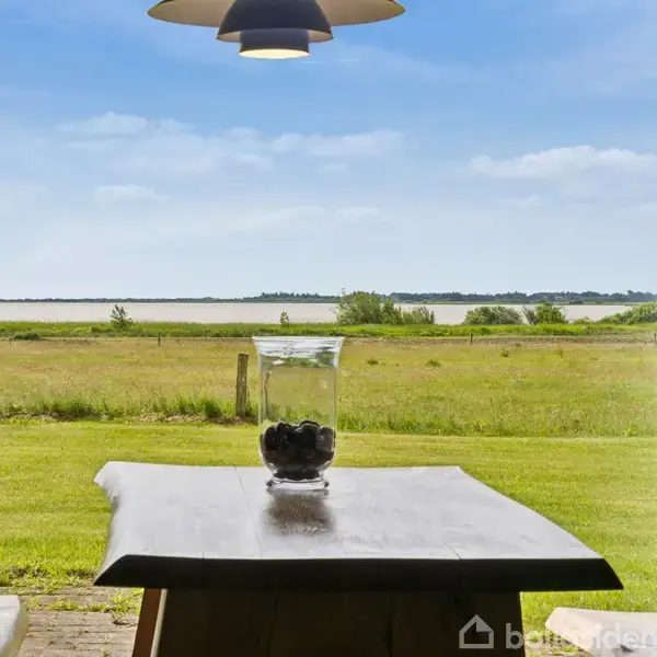 Et glas med kugler står på et træbord. Udsigt over en grøn mark og blå sø under en klar himmel. En lampe hænger over bordet.