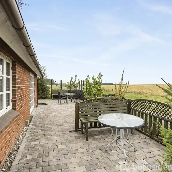 Et terrasseområde med bord og bænke langs en murstensbygning. Omgivet af landligt landskab og marker i baggrunden, giver en følelse af åbenhed og ro.