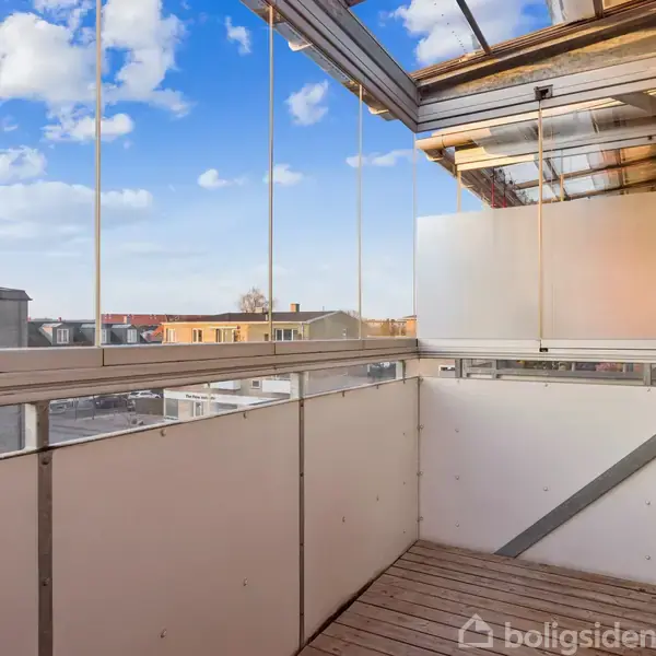 Balkon med træterrasse strækker sig ud fra en murstensbygning. Den er delvist overdækket med gennemsigtige skydedøre. I baggrunden ses byens tage under en blå himmel med skyer.