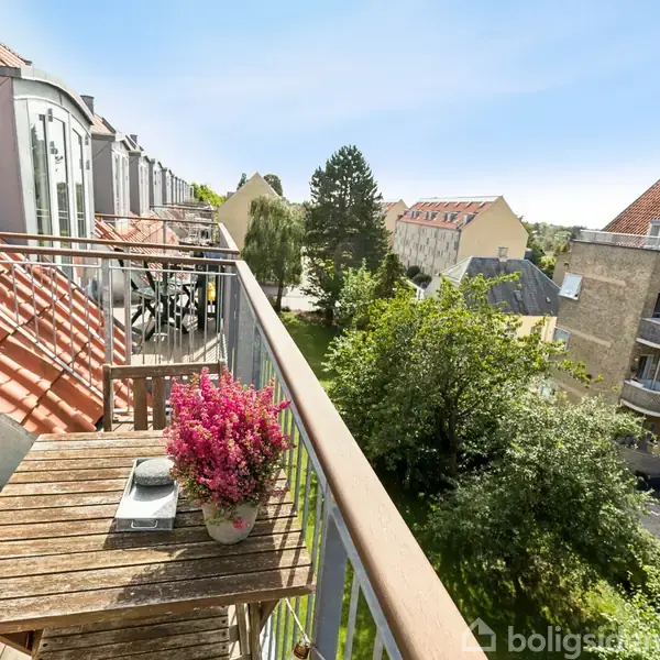 En balkon med et rustikt bord, der har en potteplante og en lille kasse ovenpå, vender ud mod en frodig have og flere boligbygninger under en klar, blå himmel.