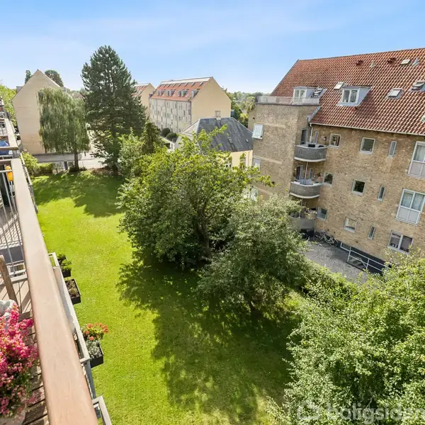 En grøn have med træer ses fra en altan, hvor blomster er placeret. I baggrunden er der fleretages bygninger med røde tage, der skaber en urban kontekst.