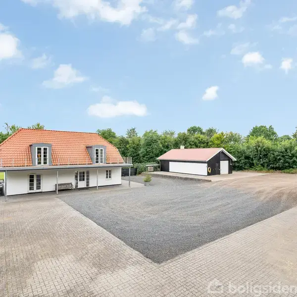 Et hvidt hus med orange tag står i en gårdsplads med grusbelægning, omgivet af grønne træer. Et mindre skur og en sort bygning er også synlige.