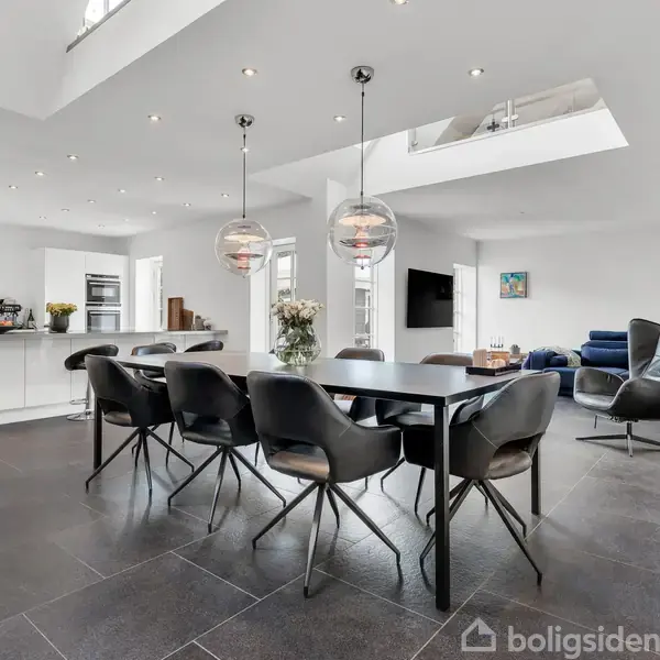 Spisebord med sorte stole står i et moderne, lyst åbent køkken-alrum. Der er to glasskuglelamper hængende over bordet og en fjernsyn i baggrunden.