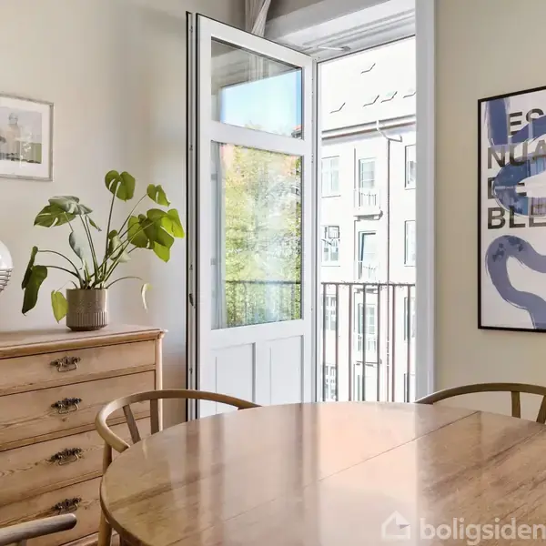 Rundt træbord foran åben dør til balkon, omgivet af træstole. Venstre væg har en kommode med plante og lampe. En plakat med teksten “ESQUISE EN BLEU Atelier Graphique A.P. 2023” hænger til højre.