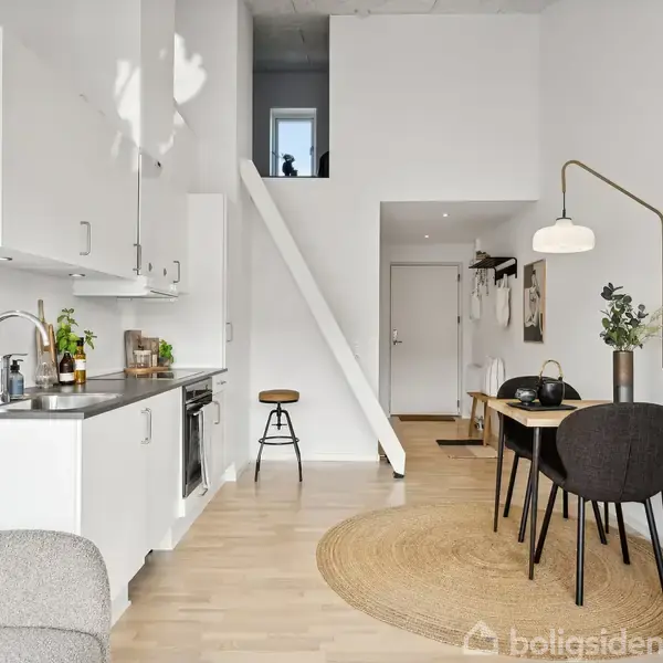 Et minimalistisk køkken med hvide skabe og integreret ovn står til venstre. Til højre er et spisebord med stole. En trappe fører til en åben hems for enden.