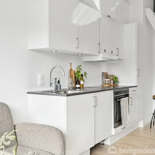 Et kompakt køkken med hvide skabe og en indbygget ovn, integreret i en moderne lys stue. Ved siden af er en skammel og en trappe fører opad.