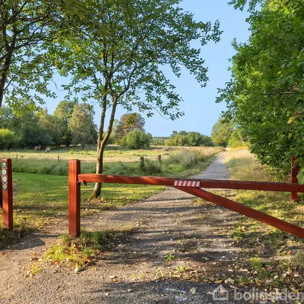 En rød træbom blokerer en grusvej i en landlig park omgivet af træer og en åben eng. En lille firkantet skilt ved bommen viser teksten "1200".