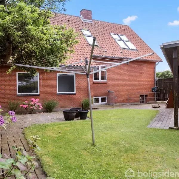 Et rødt murstenshus med græsplæne og blomster omkring. Der står en tørresnor i haven. Til højre ses en sort træbygning med terrasse. Himlen er blå med få skyer.