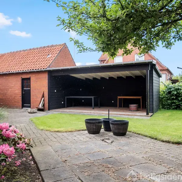 En åben garage står ved siden af et rødt murstenshus. En flisebelagt terrasse med blomstrende buske kan ses i forgrunden. Græsplænen strækker sig bagud ved siden af garagen.