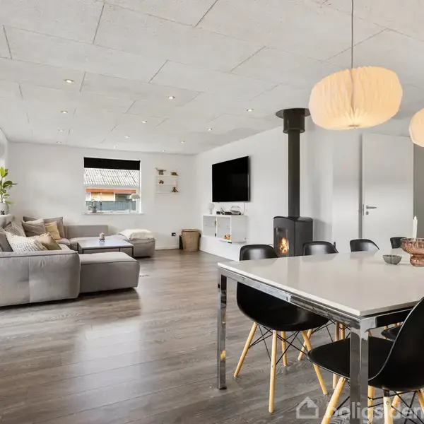 Spisebord med sorte stole står i moderne stue. Stuen har pejs, gråt sofaarrangement og væghængt tv. Store vinduer med planter skaber lys og rummelighed.