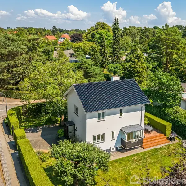 Et hvidt to-etagers hus med mørkt tag står på en grøn græsplæne omkranset af hække. Huset har en terrasse. Det er beliggende i et beboelsesområde med træer og nabohuse.