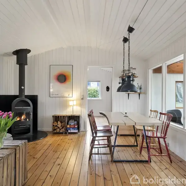 Brændeovn opvarmer hyggelig stue med trægulve; spisestue med hvide vægge og hængelys. Sofa og blomsterbord mod venstre, store vinduer til højre; kunst på væggen.