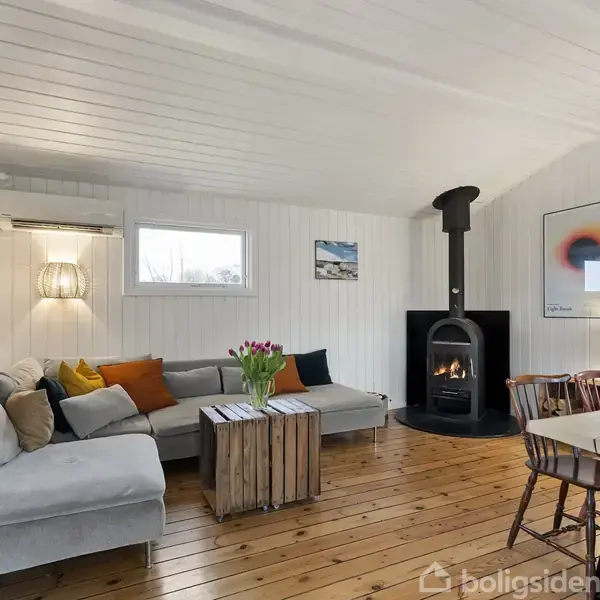 En stue med en grå sofa omkring et træbord med tulipaner, en brændeovn i et hjørne, og en spisebordssæt. Væggene er hvidmalede med kunstværker og lamper.