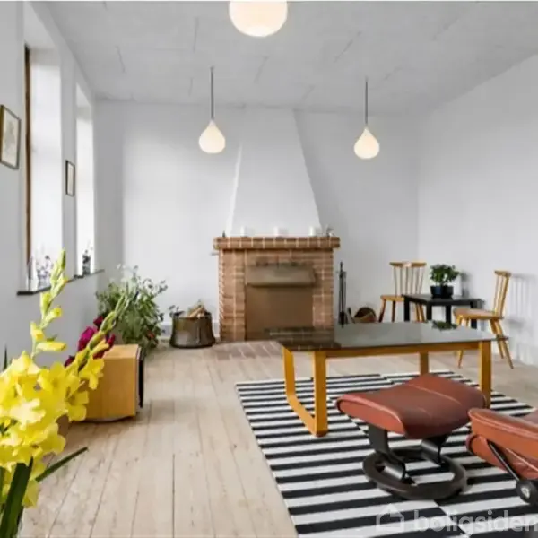 Stue med moderne indretning, hvor en blomsterdekoration står foran en pejs. Et par lænestole og et bord er placeret på et sort-hvidt stribet tæppe. Loftslamper hænger ned.