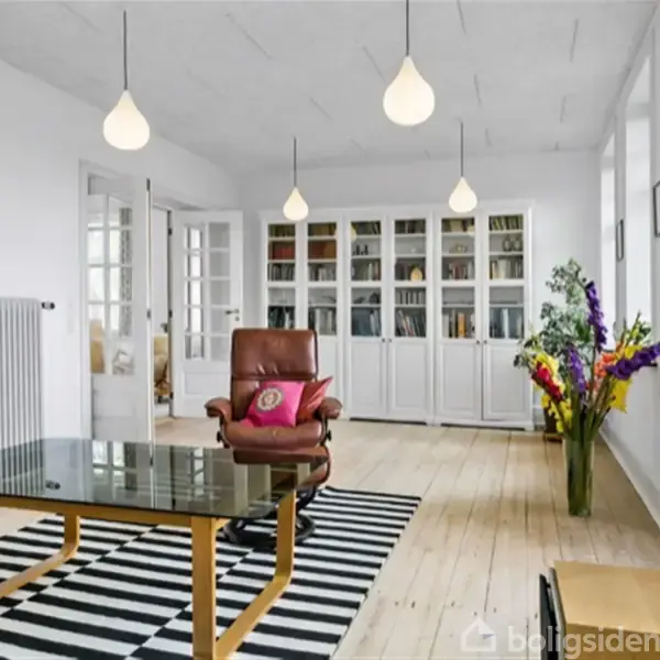 Brunt læderlænestol står ved et glasbord ovenpå et sort-hvid-stribet tæppe. Reol med bøger og blomster i vase pryder det lyse rum, oplyst af hængelamper.