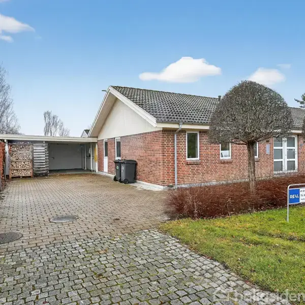 Et rødt murstenshus med carport står stille på en brostensbelagt indkørsel omgivet af trimmede træer og græsplæne. Et skilt ved indkørslen har teksten "Til Salg" med yderligere detaljer.