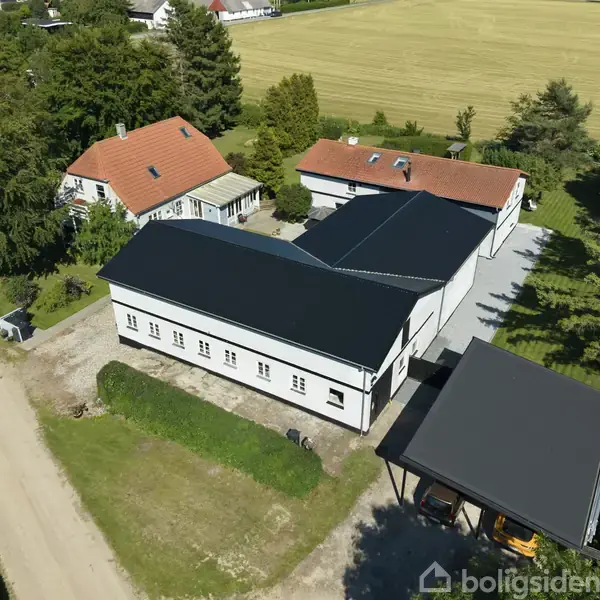 Bygning med sort tag ved en grusvej ligger i landligt område omgivet af marker og træer. Ved siden af er der en carport og et mindre hus med rødt tag.