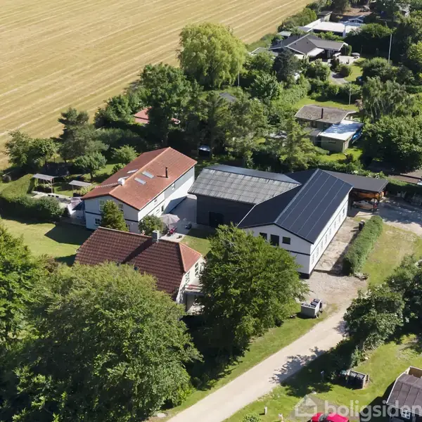 En hvid bolig med rødt tag og solpaneler står mellem grønne træer i et landskabeligt boligområde. Ved siden af ligger et stort markområde, sandsynligvis en kornmark.