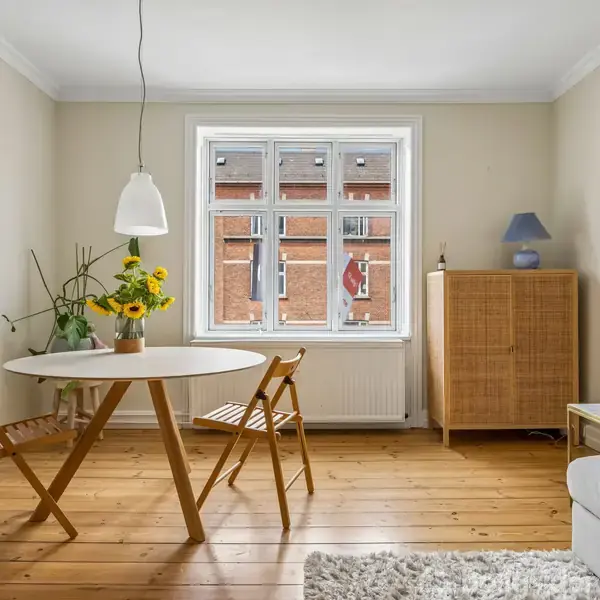 Rundt bord med blomster og to stole ved vindue i lys stue. Skænk med lampe og plakat på væggen: "SMK Schlossmuseum Charlotte von Poehl 24/6-13.8.17."